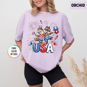 May include: A light purple t-shirt featuring a graphic of two cartoon chipmunks celebrating with fireworks and the word "USA". The chipmunks are dressed in patriotic outfits. The shirt has short sleeves and a round neckline.