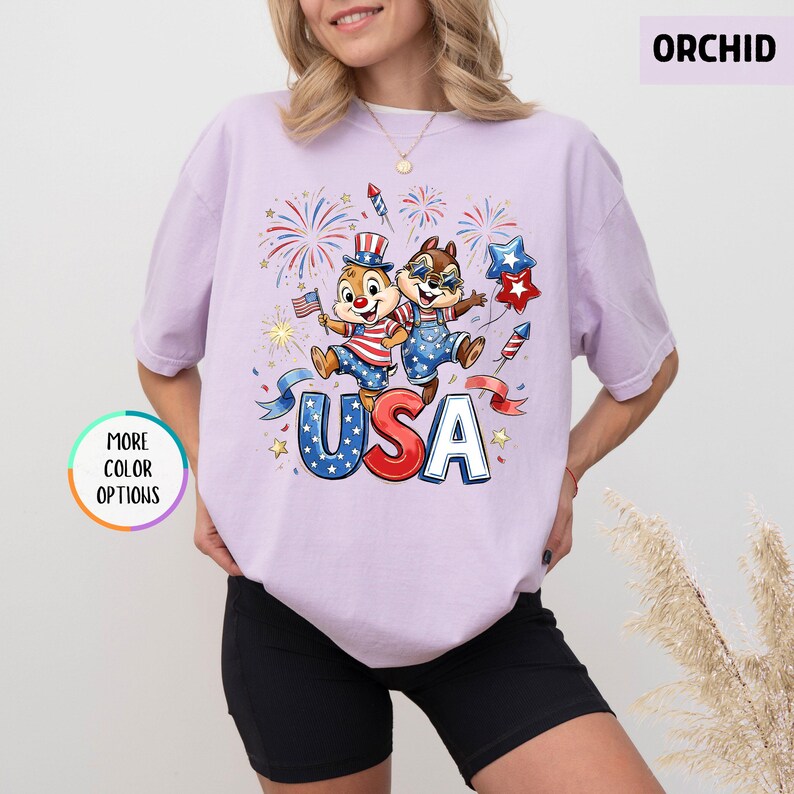 May include: A light purple t-shirt featuring a graphic of two cartoon chipmunks celebrating with fireworks and the word "USA". The chipmunks are dressed in patriotic outfits. The shirt has short sleeves and a round neckline.