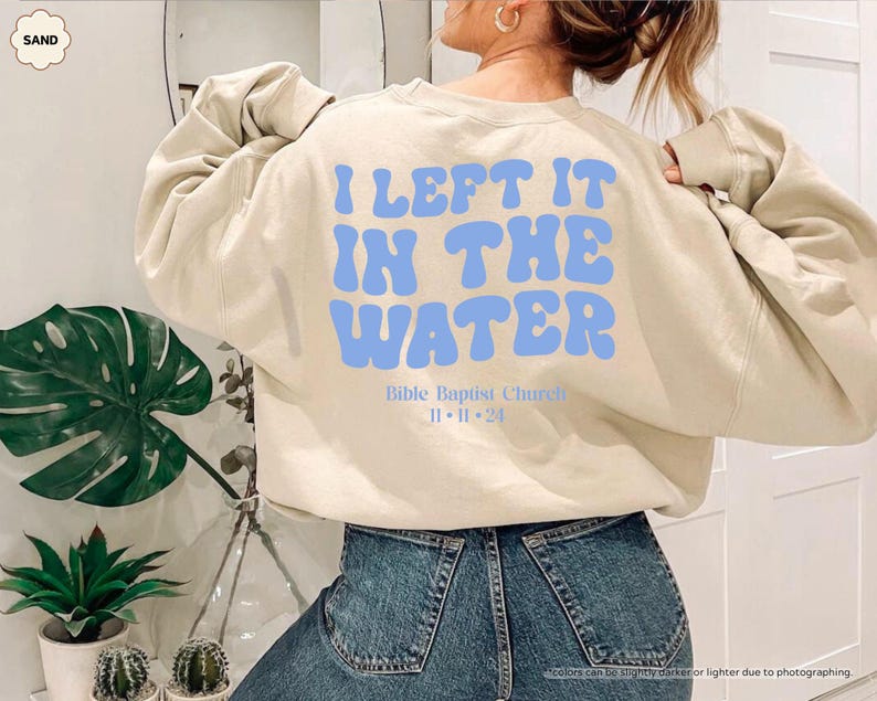 May include: Sand-colored sweatshirt with the text "I LEFT IT IN THE WATER" in light blue. Below the text is "Bible Baptist Church" and the date "11.11.24". The sweatshirt is worn by a person wearing blue jeans.