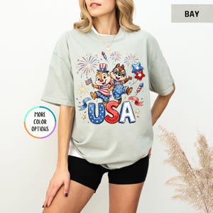 May include: A light green t-shirt featuring a graphic of two cartoon chipmunks celebrating with fireworks and the word "USA" in red, white, and blue. The shirt has short sleeves and a relaxed fit.