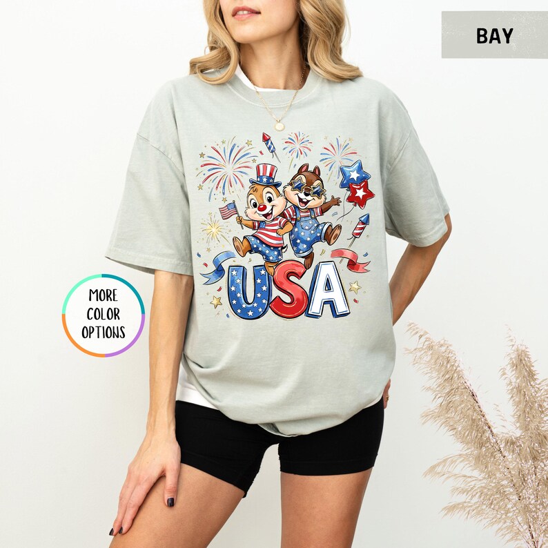May include: A light green t-shirt featuring a graphic of two cartoon chipmunks celebrating with fireworks and the word "USA" in red, white, and blue. The shirt has short sleeves and a relaxed fit.