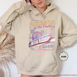 May include: Sand-colored hoodie featuring a graphic of a monorail, a sphere, and the text "PLEASE STAND CLEAR of the Doors." The design includes fireworks and text in Spanish. The hoodie has a drawstring hood and a front pocket.