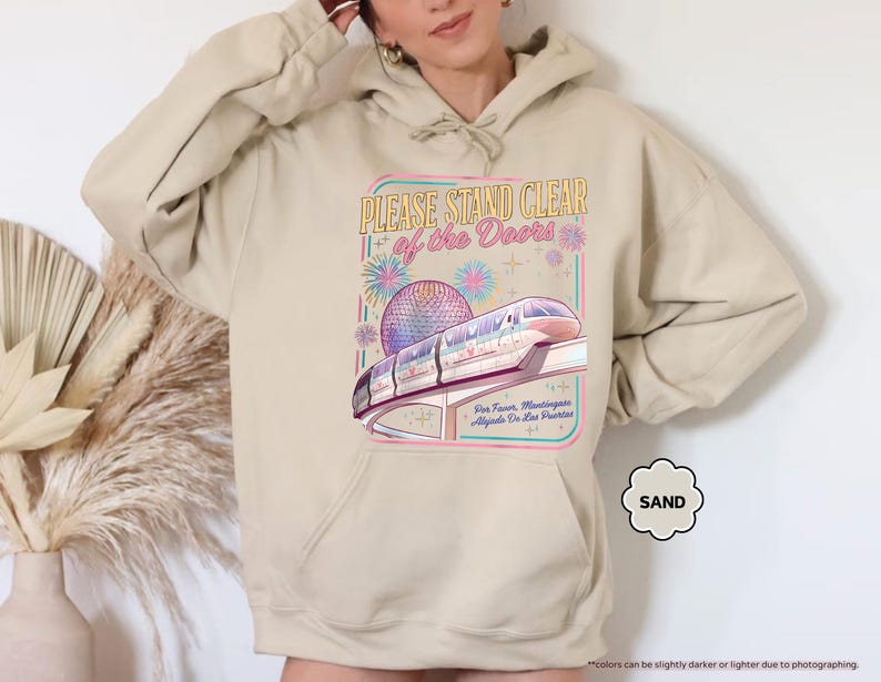 May include: Sand-colored hoodie featuring a graphic of a monorail, a sphere, and the text "PLEASE STAND CLEAR of the Doors." The design includes fireworks and text in Spanish. The hoodie has a drawstring hood and a front pocket.