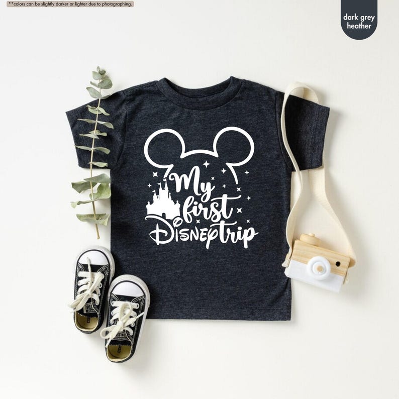 Pu&ograve; includere: Maglietta color grigio scuro melange con una grafica bianca di orecchie di Topolino, un castello e il testo "My first Disneptrip".
