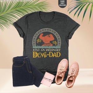 Puede incluir: Una camiseta gris oscuro jaspeada con una imagen de dibujos animados de un hombre musculoso con un cuerpo rojo y un taparrabos amarillo. El hombre est&aacute; de pie en un c&iacute;rculo con un borde amarillo y un oc&eacute;ano azul debajo. El texto "Just an ordinary Demi-Dad" est&aacute; impreso en amarillo debajo de la imagen.