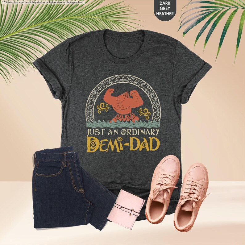 Puede incluir: Una camiseta gris oscuro jaspeada con una imagen de dibujos animados de un hombre musculoso con un cuerpo rojo y un taparrabos amarillo. El hombre est&aacute; de pie en un c&iacute;rculo con un borde amarillo y un oc&eacute;ano azul debajo. El texto "Just an ordinary Demi-Dad" est&aacute; impreso en amarillo debajo de la imagen.