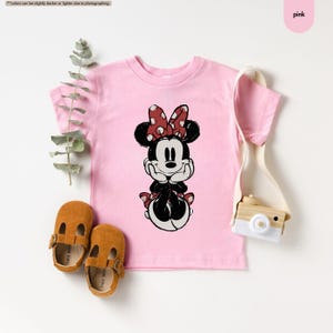 May include: A pink t-shirt featuring a Minnie Mouse graphic. The cartoon character is depicted in black, white, and red, with a polka-dot bow. The shirt is paired with brown leather baby shoes and a cream-colored bag.