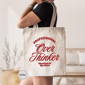 Pu&ograve; includere: Una borsa tote in tela naturale con il testo rosso "PROFESSIONAL Over Thinker Meetings In My Head". La borsa ha lunghe cinghie color crema. Una persona tiene la borsa. &Egrave; visibile una piccola etichetta circolare "NATURAL".