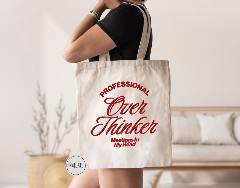 Pu&ograve; includere: Una borsa tote in tela naturale con il testo rosso "PROFESSIONAL Over Thinker Meetings In My Head". La borsa ha lunghe cinghie color crema. Una persona tiene la borsa. &Egrave; visibile una piccola etichetta circolare "NATURAL".