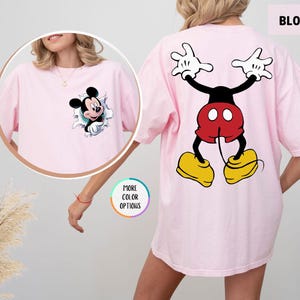 May include: A pale pink t-shirt with a Mickey Mouse design. The front has Mickey breaking through a hole, and the back shows a full cartoon image of Mickey Mouse. The word "BLOSSOM" is printed at the top right of the shirt.