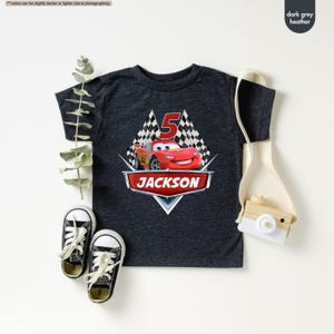 Op de afbeelding: Donkergrijs heather t-shirt met een rode raceauto-afbeelding, geblokte vlaggen en het nummer 5. De naam "JACKSON" is in rood onder de auto gedrukt.