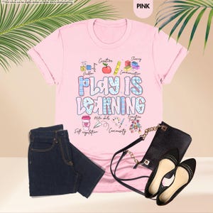 May include: A pink t-shirt with the slogan "PLAY IS LEARNING" in a blue and white checked pattern. The image includes dark blue jeans, a black handbag, and black flats. The shirt features small illustrations of a coffee cup, pencil, and paint palette.