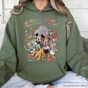 May include: Military green hoodie featuring a colorful graphic of Disney characters at the EPCOT International Flower & Garden Festival. The design includes Mickey and Minnie Mouse, Donald Duck, and Goofy, surrounded by flowers and the festival's logo.