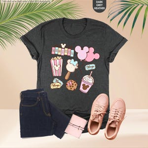 Puede incluir: Una camiseta gris oscuro jaspeada con un dise&ntilde;o caprichoso que presenta orejas de Mickey Mouse, palomitas de ma&iacute;z, un helado de Mickey Mouse, una galleta de Mickey Mouse, una bebida con tem&aacute;tica de Mickey Mouse y la palabra "MAGICAL" en letras rosas.