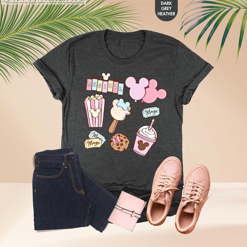 Puede incluir: Una camiseta gris oscuro jaspeada con un dise&ntilde;o caprichoso que presenta orejas de Mickey Mouse, palomitas de ma&iacute;z, un helado de Mickey Mouse, una galleta de Mickey Mouse, una bebida con tem&aacute;tica de Mickey Mouse y la palabra "MAGICAL" en letras rosas.