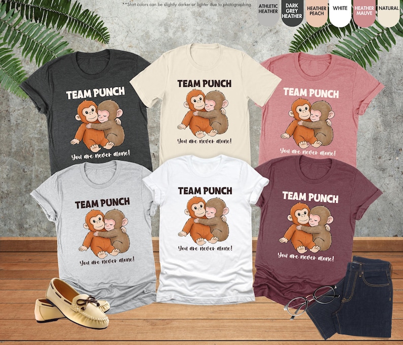 Puede incluir: Una colecci&oacute;n de camisetas en varios colores, incluyendo gris jaspeado, blanco y malva. Cada camiseta presenta un gr&aacute;fico de dos monos de dibujos animados abraz&aacute;ndose, con el texto "TEAM PUNCH" y "You are never alone!"