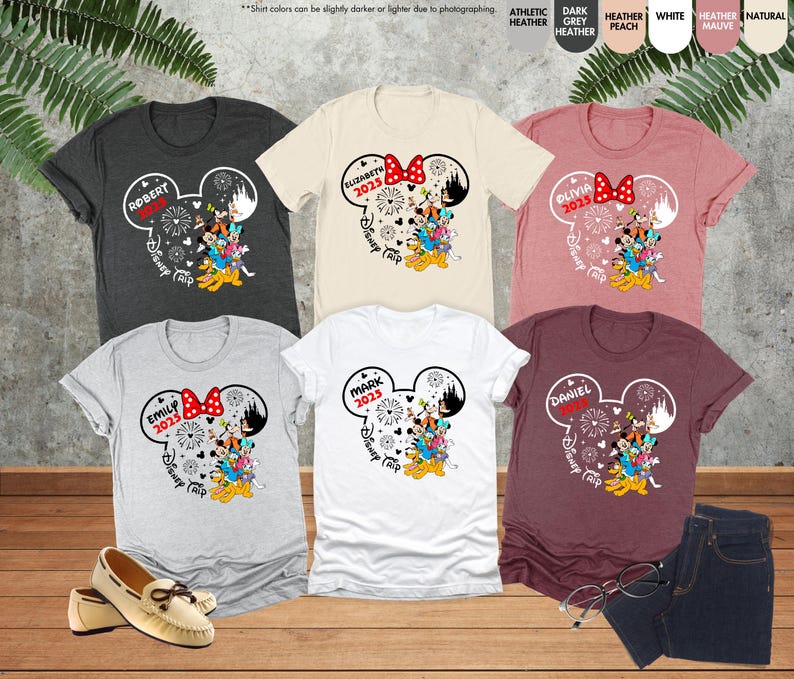 May include: Six t-shirts with a Mickey Mouse head design. Each shirt has a different name and the year 2023 printed on it. The shirts are in various colors, including gray, white, and maroon.