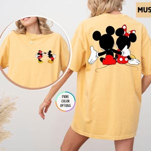 May include: Mustard yellow t-shirt with a Mickey and Minnie Mouse graphic. The front shows the characters kissing, and the back features them embracing. The shirt has a relaxed, oversized fit, suitable for casual wear.