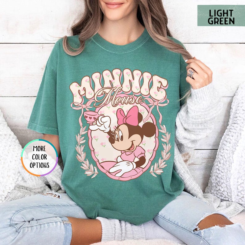 May include: A light green t-shirt with a vintage-style graphic of Minnie Mouse holding a teacup. The graphic features the text "Minnie Mouse" in a retro font.