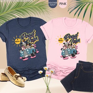 Op de afbeelding: Twee T-shirts, &eacute;&eacute;n marineblauw en &eacute;&eacute;n roze, met een "Road Trip! Miami"-afbeelding met stripfiguren in een blauwe auto. Een paar cr&egrave;mekleurige loafers en een blauwe jeans zijn ook zichtbaar.