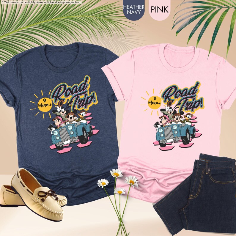 Op de afbeelding: Twee T-shirts, &eacute;&eacute;n marineblauw en &eacute;&eacute;n roze, met een "Road Trip! Miami"-afbeelding met stripfiguren in een blauwe auto. Een paar cr&egrave;mekleurige loafers en een blauwe jeans zijn ook zichtbaar.
