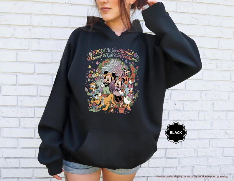 May include: Black hooded sweatshirt featuring a colorful graphic of Disney characters at the EPCOT International Flower & Garden Festival. The design includes Mickey and Minnie Mouse, Donald Duck, and Pluto, surrounded by flowers. The word "BLACK" is in a black bubble.