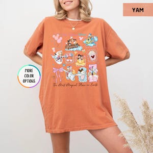 May include: A yam-colored t-shirt featuring a colorful graphic of cartoon characters and the text "The Most Magical Place on Earth." The design includes various characters and icons. The shirt has short sleeves and a relaxed fit.