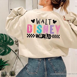 Puede incluir: Una sudadera de color arena con el texto "WALT DISNEY WORLD" en letras coloridas y un patr&oacute;n de cuadros blancos y negros. La sudadera tiene mangas largas y est&aacute; hecha de un material suave.