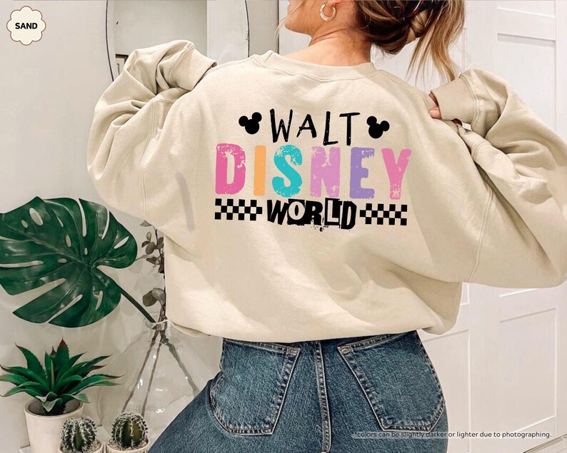 Puede incluir: Una sudadera de color arena con el texto "WALT DISNEY WORLD" en letras coloridas y un patr&oacute;n de cuadros blancos y negros. La sudadera tiene mangas largas y est&aacute; hecha de un material suave.