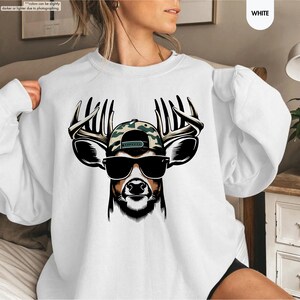 May include: White sweatshirt with a graphic of a deer wearing sunglasses and a camouflage baseball cap. The deer has large antlers.