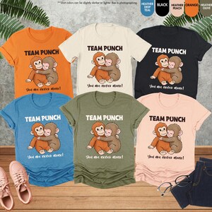 Puede incluir: Una colecci&oacute;n de camisetas en varios colores, incluyendo naranja, azul, negro y melocot&oacute;n. Cada camiseta presenta el texto "TEAM PUNCH" sobre una ilustraci&oacute;n de dos monos abraz&aacute;ndose. Debajo est&aacute; el texto "You are never alone!"