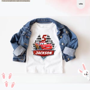 Op de afbeelding: Wit T-shirt met een rode raceauto-afbeelding, geblokte vlaggen en het nummer 5. De tekst "JACKSON" staat onder de auto.