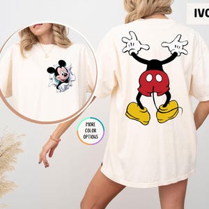 May include: Ivory t-shirt featuring Mickey Mouse. The front has a graphic of Mickey breaking through a wall, while the back displays a full-body image of Mickey Mouse. The shirt is a relaxed fit.