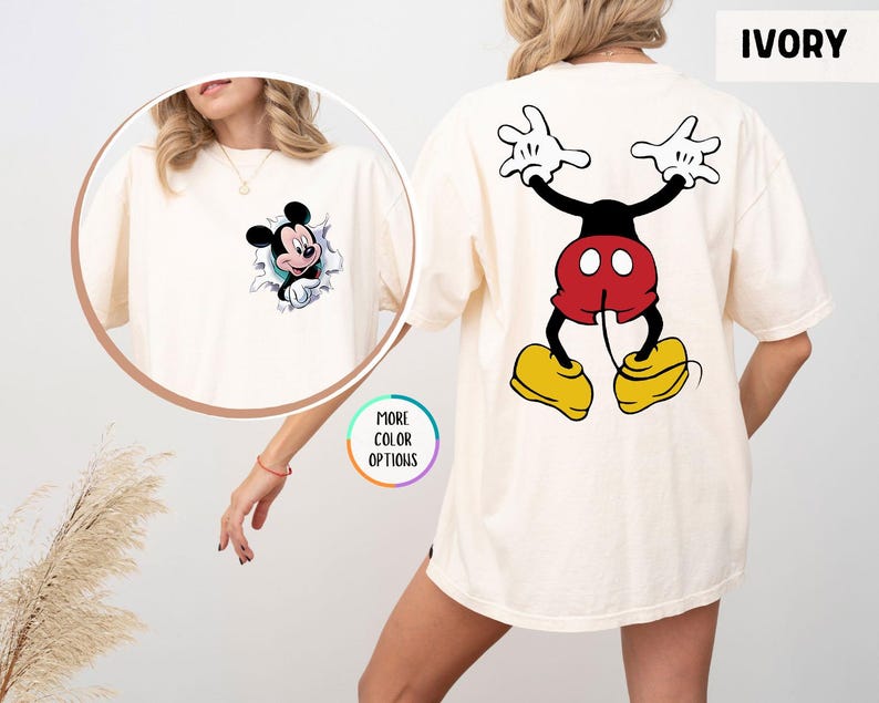 May include: Ivory t-shirt featuring Mickey Mouse. The front has a graphic of Mickey breaking through a wall, while the back displays a full-body image of Mickey Mouse. The shirt is a relaxed fit.