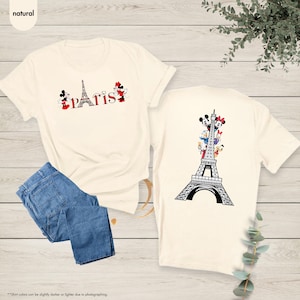 Pu&ograve; includere: T-shirt color crema con grafica "Parigi" con i personaggi di Topolino e Minnie. Il retro della maglietta mostra la Torre Eiffel con i personaggi Disney. Jeans blu piegati accanto alla maglietta.
