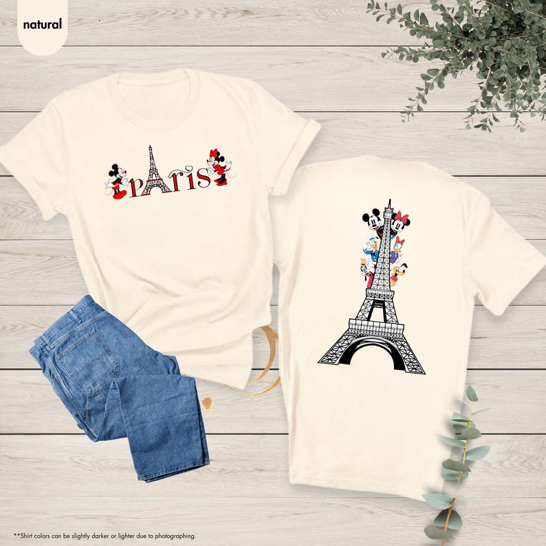 Pu&ograve; includere: T-shirt color crema con grafica "Parigi" con i personaggi di Topolino e Minnie. Il retro della maglietta mostra la Torre Eiffel con i personaggi Disney. Jeans blu piegati accanto alla maglietta.