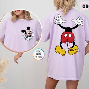 May include: Orchid-coloured t-shirt with a Mickey Mouse graphic. The front shows Mickey breaking through a torn paper effect, and the back displays a full Mickey Mouse design. The shirt has short sleeves and a relaxed fit. The word "ORCHID" is visible.