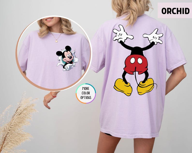 May include: Orchid-coloured t-shirt with a Mickey Mouse graphic. The front shows Mickey breaking through a torn paper effect, and the back displays a full Mickey Mouse design. The shirt has short sleeves and a relaxed fit. The word "ORCHID" is visible.