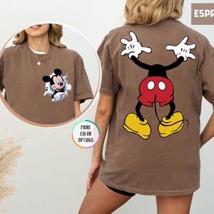 May include: Espresso-coloured t-shirt featuring Mickey Mouse. The front has a small Mickey graphic bursting through a hole. The back displays a larger Mickey Mouse design with white gloves, red shorts, and yellow shoes. "MORE COLOR OPTIONS" is also visible.