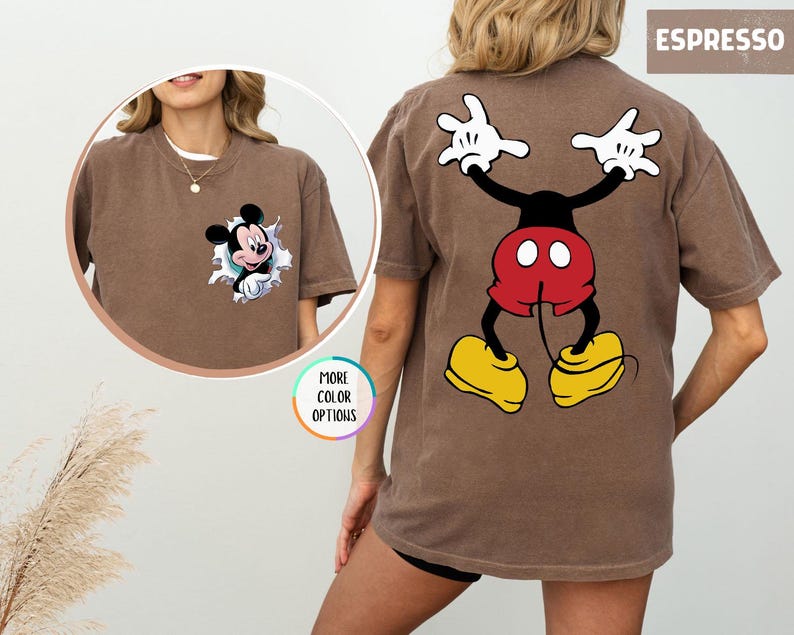 May include: Espresso-coloured t-shirt featuring Mickey Mouse. The front has a small Mickey graphic bursting through a hole. The back displays a larger Mickey Mouse design with white gloves, red shorts, and yellow shoes. "MORE COLOR OPTIONS" is also visible.