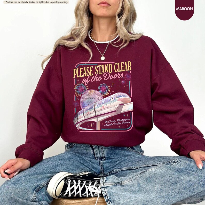 May include: Maroon sweatshirt with the text "PLEASE STAND CLEAR of the Doors" and a graphic of a monorail and fireworks. The sweatshirt is a solid maroon color and is made of a soft material.