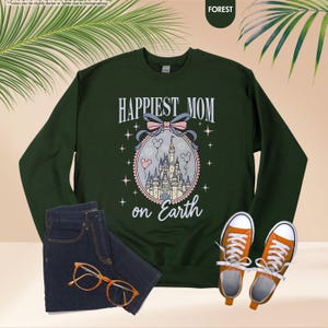 May include: A forest green sweatshirt with the text "HAPPIEST MOM on Earth" above a castle illustration. The image includes a pair of blue jeans, orange sneakers, and eyeglasses.