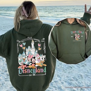 May include: A green hooded sweatshirt with a graphic print of Mickey Mouse and friends in front of the Disneyland castle. The text "Disneyland Est. 1955" is printed below the graphic. The sweatshirt is shown on a model standing on a beach.