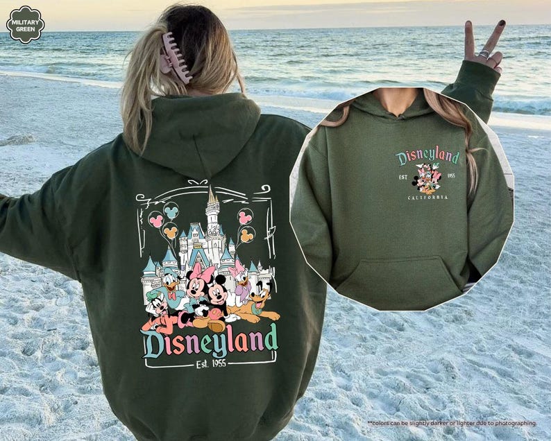 May include: A green hooded sweatshirt with a graphic print of Mickey Mouse and friends in front of the Disneyland castle. The text "Disneyland Est. 1955" is printed below the graphic. The sweatshirt is shown on a model standing on a beach.