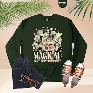 Pode incluir: Camisola verde floresta com um gr&aacute;fico colorido que diz "The Most Magical Place". O gr&aacute;fico inclui um castelo, Mickey Mouse e outros elementos do parque tem&aacute;tico. Combinada com jeans azuis, t&ecirc;nis laranja e &oacute;culos.