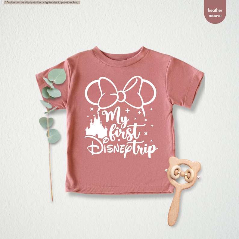 Pu&ograve; includere: Una maglietta color malva con un design grafico bianco che dice "My first Disneptrip" con una silhouette bianca del castello di Cenerentola e un fiocco bianco con orecchie da topo.