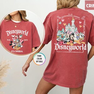 Puede incluir: Camiseta de color carmes&iacute; con un dise&ntilde;o de Disneyworld. La parte delantera presenta a Mickey, Minnie, Donald y otros personajes con el texto "Disneyworld Florida Est. 1971." La parte trasera muestra un castillo y personajes con "Disneyworld 1971."