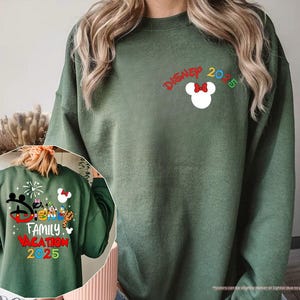K&ouml;nnte beinhalten: Ein gr&uuml;ner Pullover mit dem Text "DISNEY 2025" und einem Mickey Mouse Kopf mit einer roten Schleife. Der Pullover zeigt auch eine kleinere Grafik von Mickey Mouse und anderen Disney-Charakteren mit dem Text "FAMILY VACATION 2025".
