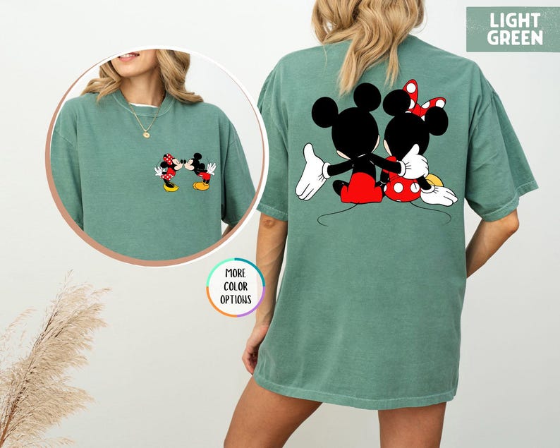 May include: Light green t-shirt with Mickey and Minnie Mouse graphics. A small image of the characters is on the front, and a larger image of them embracing is on the back. The shirt has a relaxed, oversized fit.