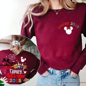 K&ouml;nnte beinhalten: Ein maroonfarbener Pullover mit dem Text "DISNEY 2025" und einem Cartoon-Bild von Mickeys Kopf. Die R&uuml;ckseite des Pullovers zeigt eine bunte Grafik von Mickey Mouse und anderen Disney-Charakteren mit dem Text "DISNEY FAMILY VACATION 2025".